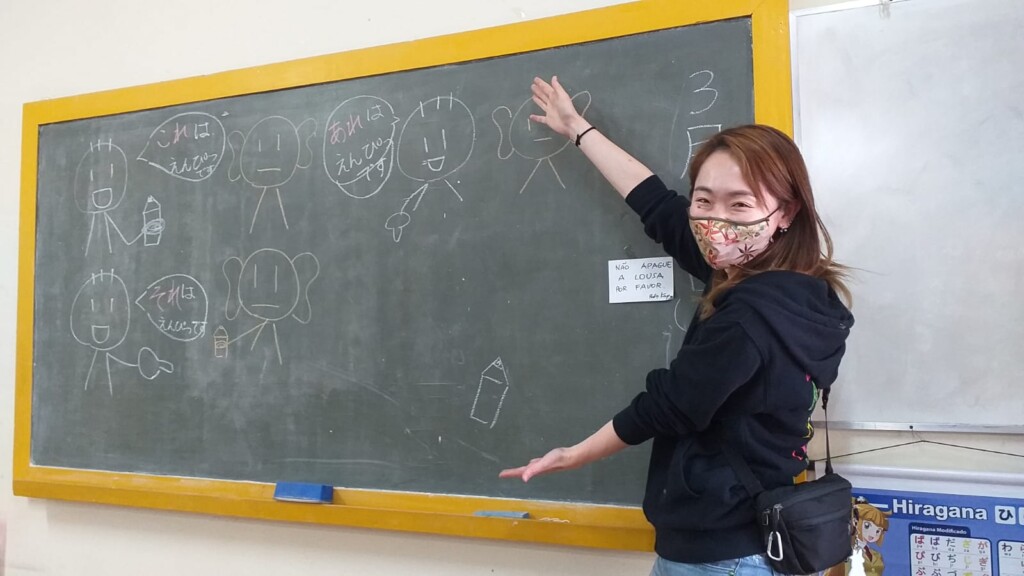 ２年前そのままの黒板を前にした櫻井さん。右下に「Não apague a lousa, por favor（黒板を消さないで）」と張り紙（本人提供）