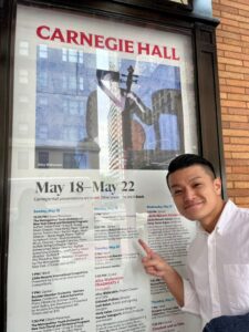 「銀座男声合唱団」が記載されたカーネギーホールのタイムテーブルと記念撮影する本田氏